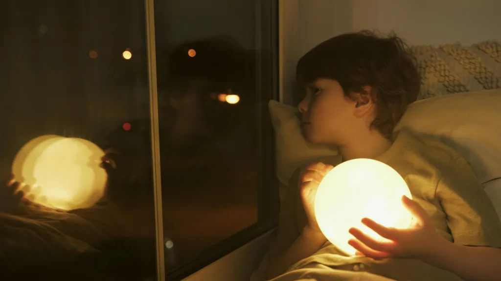 Un enfant dans son lit, tient une veilleuse dans ses mains et regarde par la fenêtre
